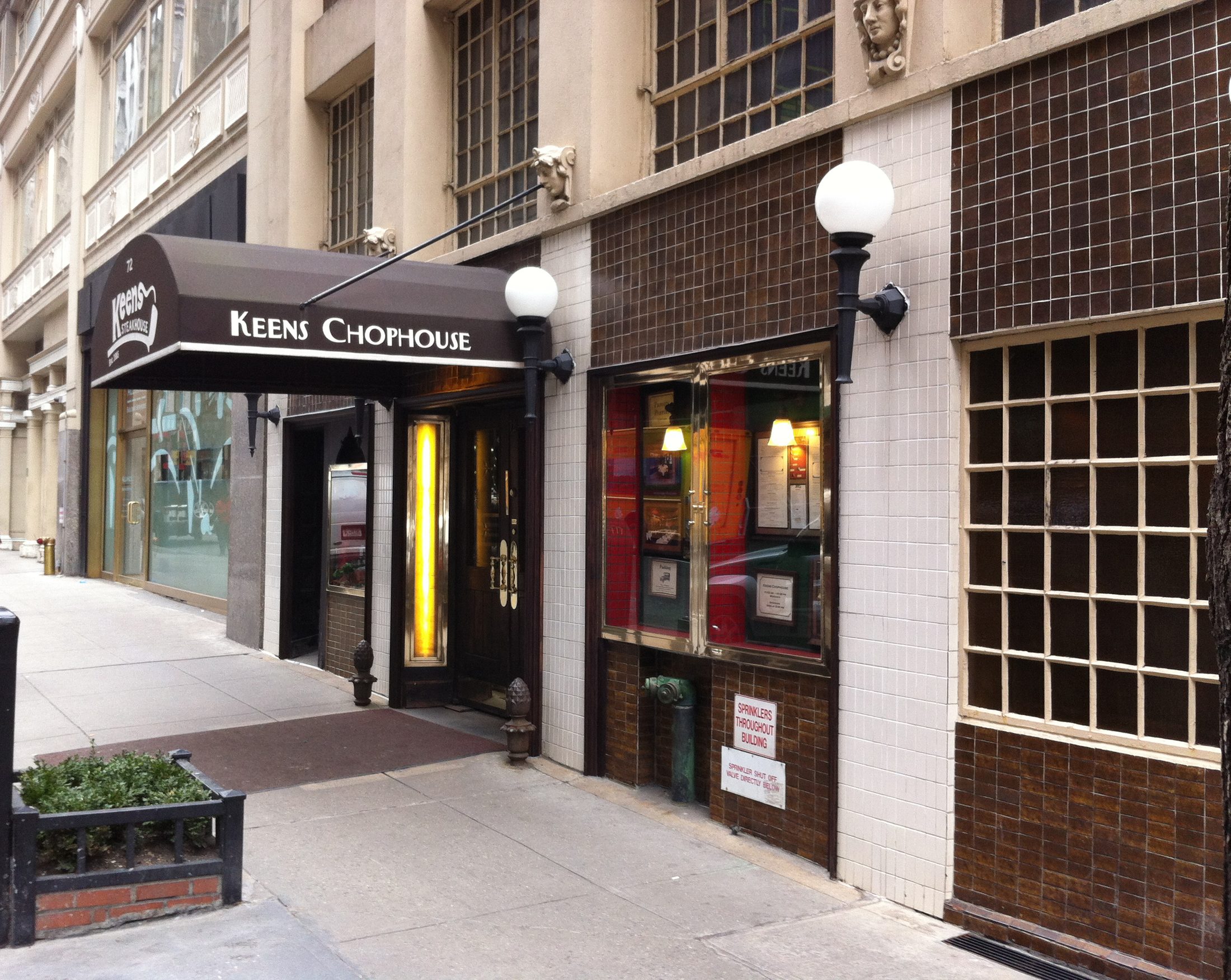 The Keens Chophouse awning and globe lamps over the dark wooden entrance of Keens Steakhouse on a quiet Midtown sidewalk.