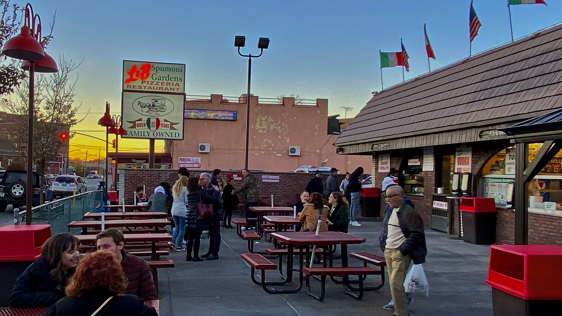 Diners at the outdoor picnic tables of L&B Spumoni Gardens on 86th Street in Gravesend, Brooklyn, under its pizzeria sign at dusk
