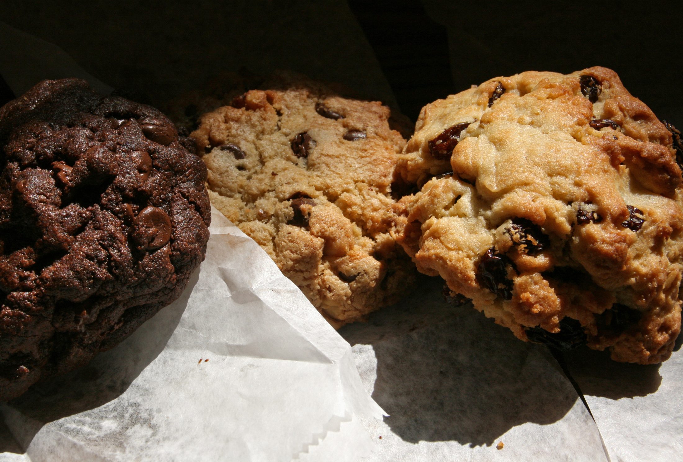 Three craggy, oversized Levain Bakery cookies — chocolate chocolate chip, oatmeal raisin and chocolate chip walnut — on wax paper.