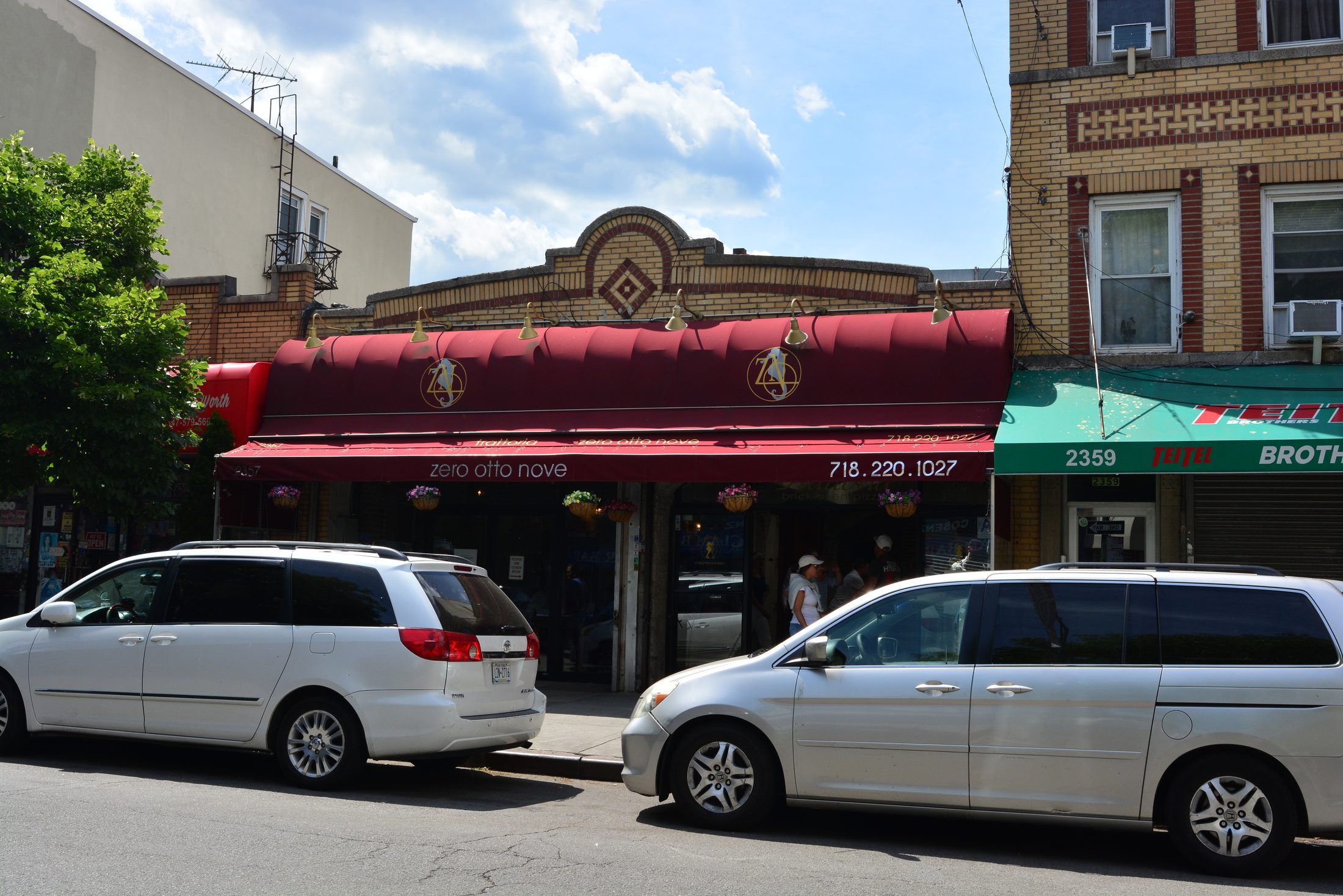 Zero Otto Nove's red awning and brick facade on Arthur Avenue in the Bronx, with minivans parked at the curb.
