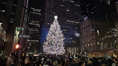 The Rockefeller Center Christmas tree lit at night above a crowd, with the tower of 30 Rockefeller Plaza behind